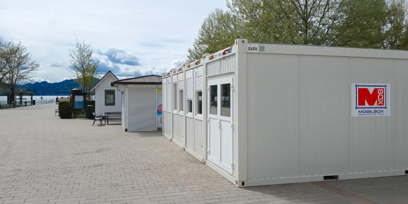 Ein Mobilbox Bürocontainer von außen, platziert am Ufer des Wörthersees in Klagenfurt. Die Container haben Fenster mit geschlossenen Rollläden und eine Eingangstür. Das Mobilbox-Logo ist an den Containern sichtbar. Im Hintergrund sind der See und eine bergige Landschaft zu erkennen.